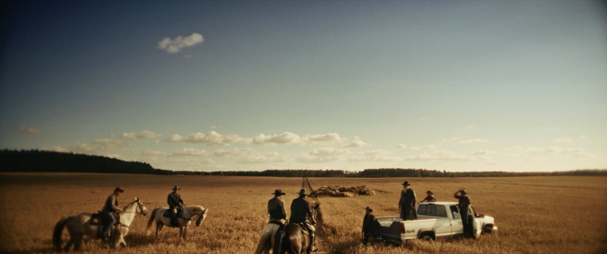 Riders on horseback and a pickup gathered at the edge of a vast golden field under a blue sky - Amon Amarth